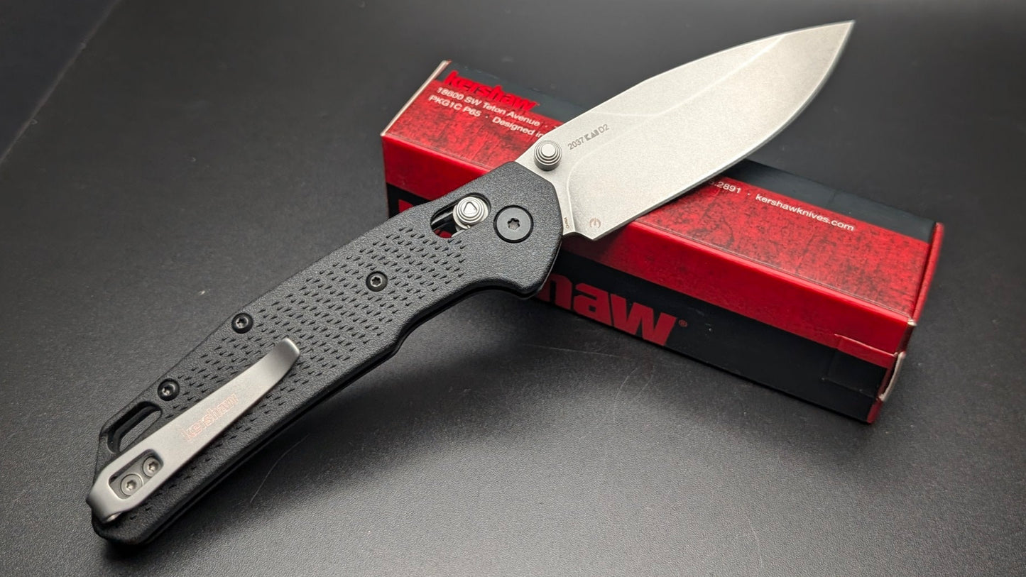 Folding knife with a black handle and silver blade on a dark surface with a red box featuring 'Kershaw' branding.