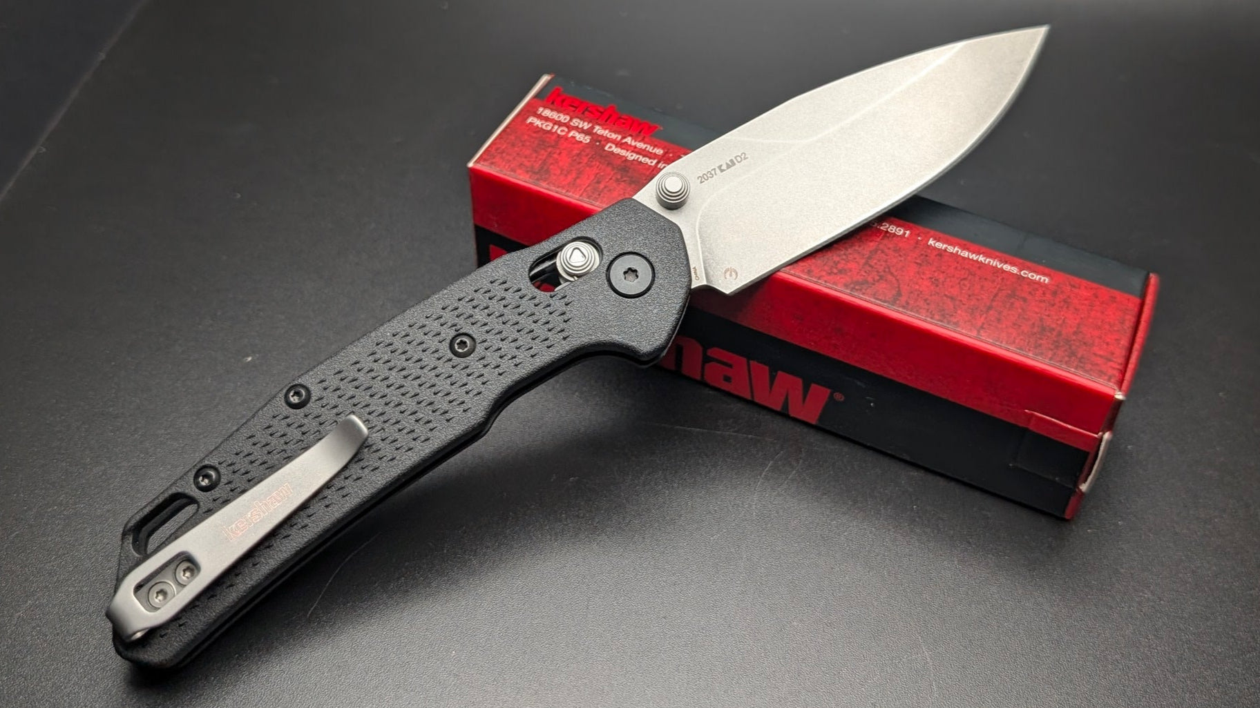 Folding knife with a black handle and silver blade on a dark surface with a red box featuring 'Kershaw' branding.