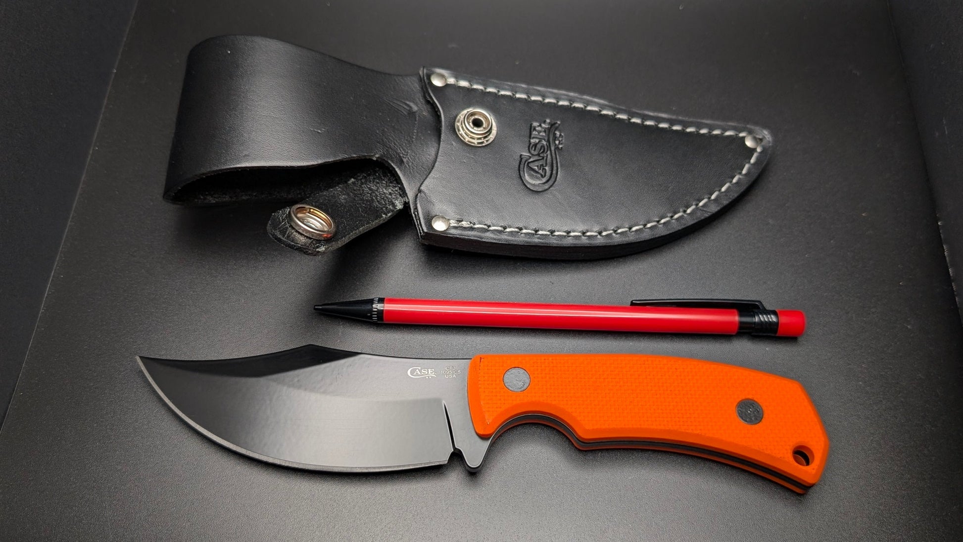 Knife with orange handle and black blade, pen, and leather sheath on a dark surface