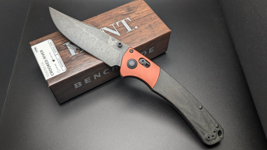 Benchmade knife with brown handle and gray blade on a dark surface with a Hunt box in the background.