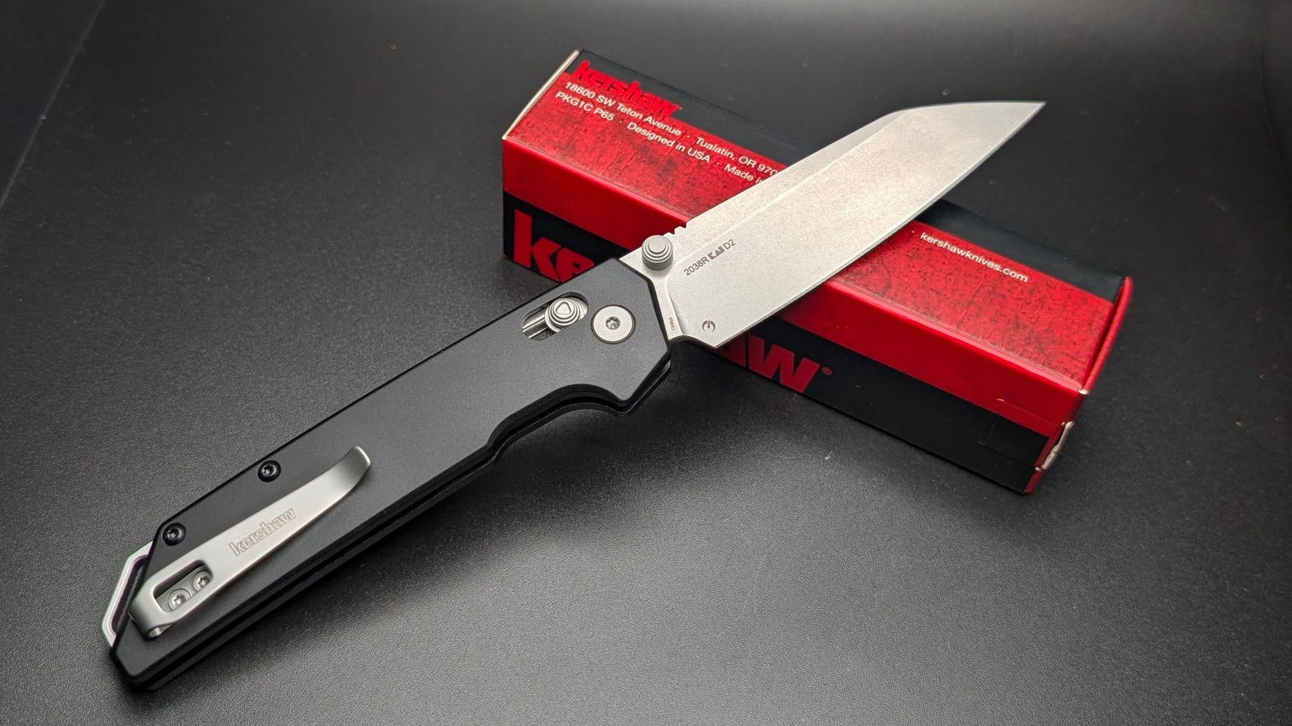 Folding knife with a black handle and silver blade on a dark surface, accompanied by a red and black box.